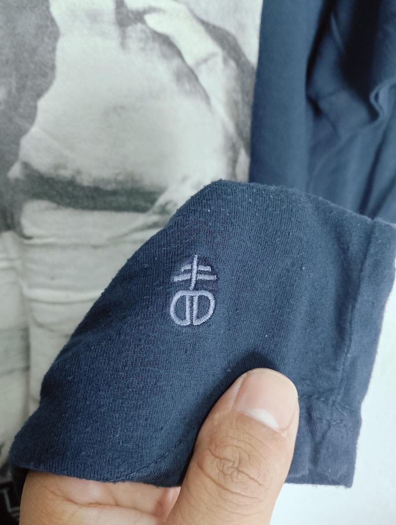 May include: Close-up of a navy blue long-sleeved shirt. The shirt features a light gray embroidered logo on the sleeve. The background shows a black and white graphic print.