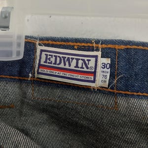 Puede incluir: Primer plano de tejido vaquero azul con una etiqueta "EDWIN" en letras blancas sobre fondo morado. La etiqueta indica "SOLD ONLY AT THE FINEST STORES". Indica una cintura de 30 pulgadas (76 cm) y una longitud de 76 cm.