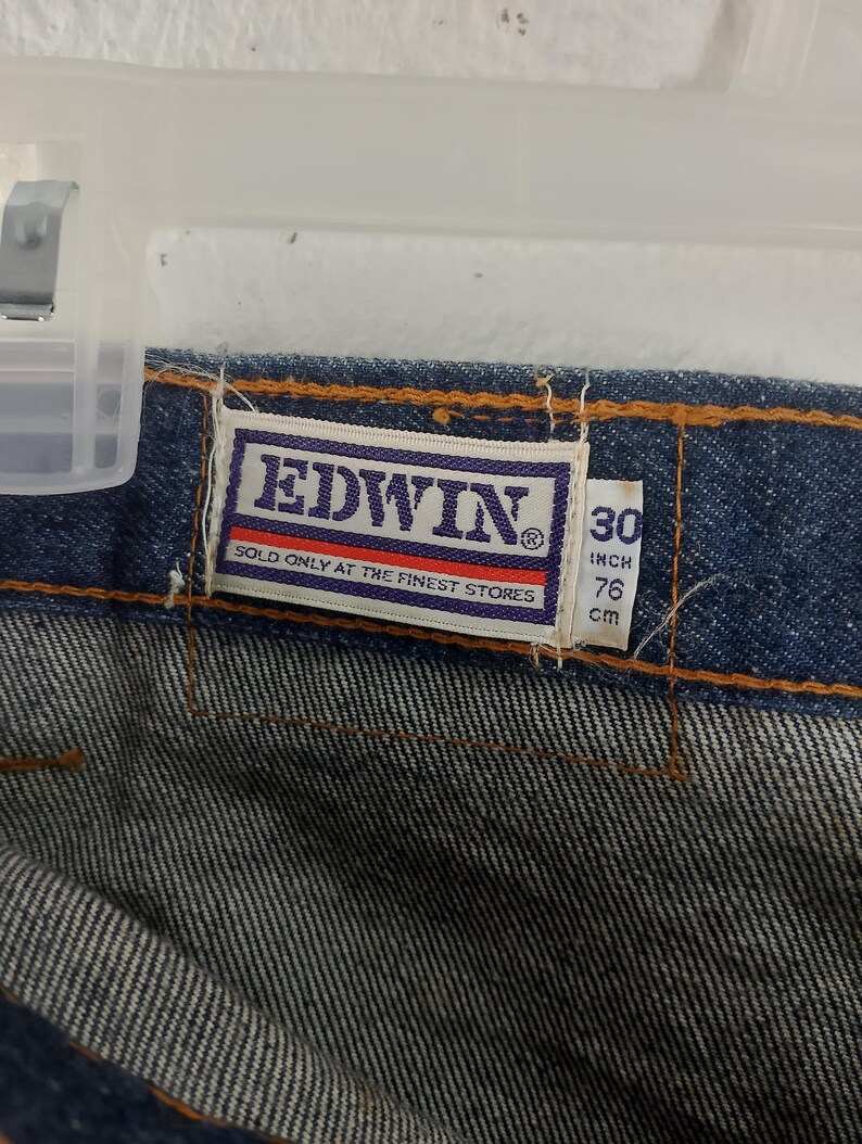 Puede incluir: Primer plano de tejido vaquero azul con una etiqueta "EDWIN" en letras blancas sobre fondo morado. La etiqueta indica "SOLD ONLY AT THE FINEST STORES". Indica una cintura de 30 pulgadas (76 cm) y una longitud de 76 cm.