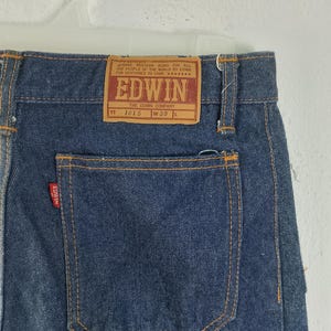 Puede incluir: Primer plano de la parte trasera de unos vaqueros azules con una etiqueta rectangular marr&oacute;n que dice "EDWIN" y "THE EDWIN COMPANY". Los vaqueros tienen un bolsillo trasero y son de la talla W30 L.