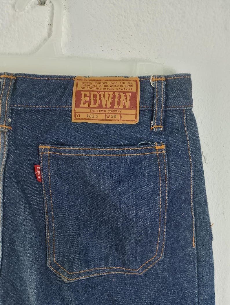 Puede incluir: Primer plano de la parte trasera de unos vaqueros azules con una etiqueta rectangular marr&oacute;n que dice "EDWIN" y "THE EDWIN COMPANY". Los vaqueros tienen un bolsillo trasero y son de la talla W30 L.