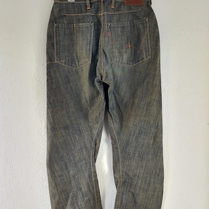 Può includere: Vista posteriore di jeans in denim dall'aspetto sbiadito e strutturato. I jeans presentano due tasche posteriori, cuciture arancioni e una toppa in pelle marrone. I jeans sono appesi a una gruccia bianca.