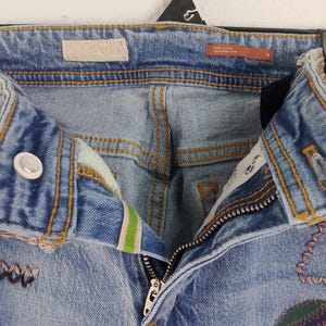 Pode incluir: Jeans de ganga azul claro com aplica&ccedil;&otilde;es decorativas e costuras coloridas. Os jeans apresentam fecho de bot&atilde;o, z&iacute;per e uma etiqueta que diz "made in china". Os jeans s&atilde;o tamanho 26.