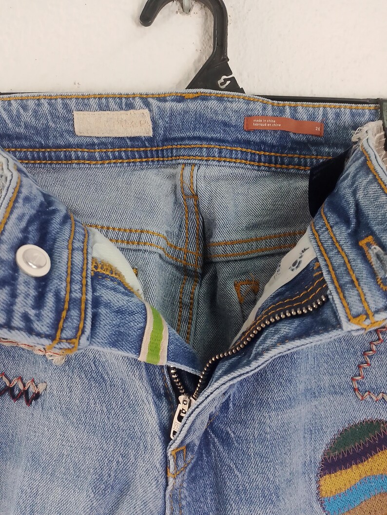 Pode incluir: Jeans de ganga azul claro com aplica&ccedil;&otilde;es decorativas e costuras coloridas. Os jeans apresentam fecho de bot&atilde;o, z&iacute;per e uma etiqueta que diz "made in china". Os jeans s&atilde;o tamanho 26.
