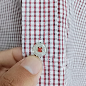 K&ouml;nnte beinhalten: Ein wei&szlig;es Hemd mit einem kleinen rot-dunkelbraunen Vichy-Karomuster. Das Hemd verf&uuml;gt &uuml;ber eine mittige Knopfleiste mit einem runden, durchscheinenden Knopf, der mit dem Text Ben Sherman graviert und mit rotem Kreuzstichgarn befestigt ist.