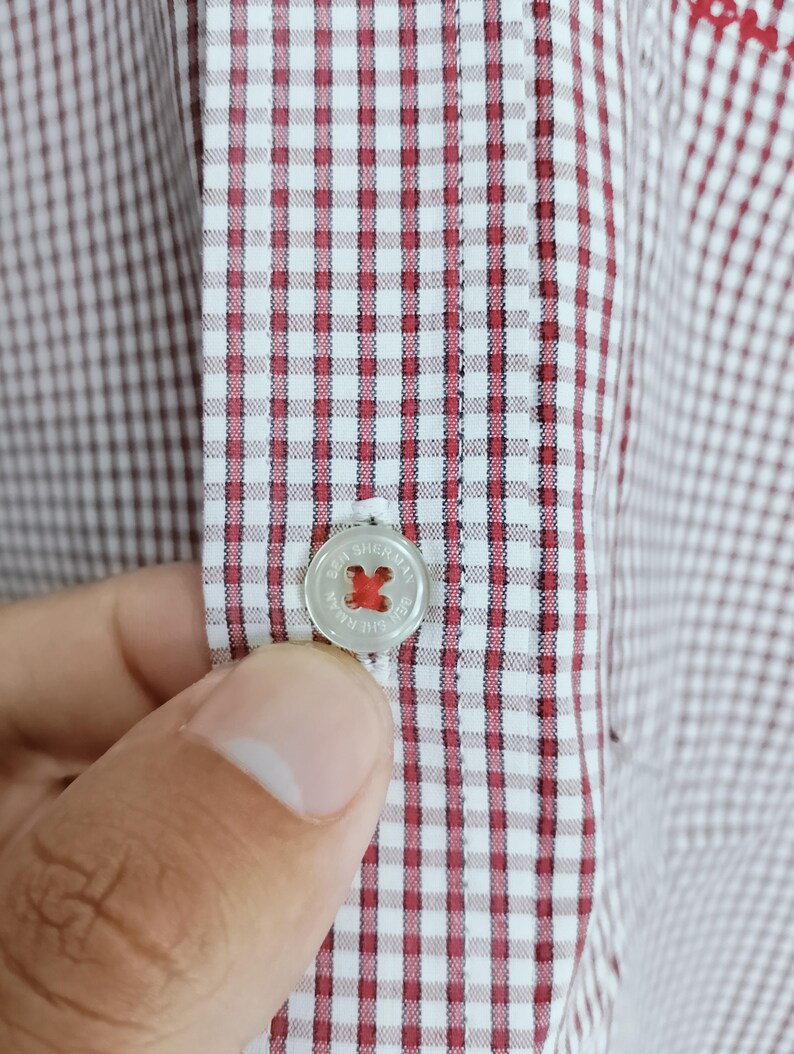 K&ouml;nnte beinhalten: Ein wei&szlig;es Hemd mit einem kleinen rot-dunkelbraunen Vichy-Karomuster. Das Hemd verf&uuml;gt &uuml;ber eine mittige Knopfleiste mit einem runden, durchscheinenden Knopf, der mit dem Text Ben Sherman graviert und mit rotem Kreuzstichgarn befestigt ist.