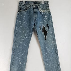 Puede incluir: Vaqueros de mezclilla azul claro con detalles desgastados, que incluyen rasgaduras y salpicaduras de pintura blanca. Los jeans presentan un diseño de rayo negro recortado en la pierna derecha. Los jeans están colgados en una percha blanca.