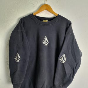 May include: Dark navy blue sweatshirt with white diamond-shaped logo on the chest and sleeves. The sweatshirt is hanging on a wooden hanger against a white wall. The brand name is visible on the collar.