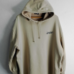May include: A light beige hooded sweatshirt with a black graphic of the word "Arvail" on the front.