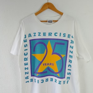 May include: White t-shirt with a colorful graphic celebrating 25 years of Jazzercise. The design features a teal square with the words "JAZZERCISE" in blue, surrounding a large yellow star and the number "25" in purple.