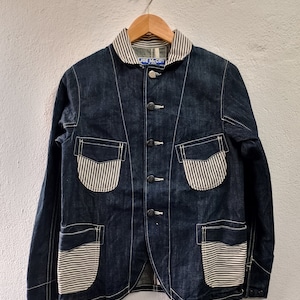 May include: A dark blue denim jacket with a white and black striped collar and pocket trim. The jacket has a button closure and multiple pockets. The label on the jacket reads "JOE McCOY".