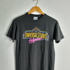 Może przedstawiać: Czarny t-shirt z logo Universal Studios Hollywood w neonowych kolorach. Logo zawiera napisy "Universal Studios" nad "Hollywood" w kursywie. Koszulka wisi na drewnianym wieszaku.