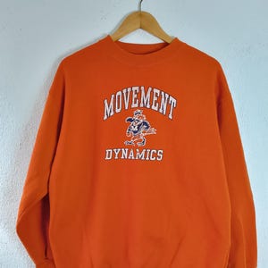 以下が含まれることがあります： オレンジ色のスウェットシャツ。白文字で「MOVEMENT DYNAMICS」と書かれています。「MOVEMENT」の下には漫画の虎のグラフィックがあります。スウェットシャツは木製のハンガーに吊るされています。