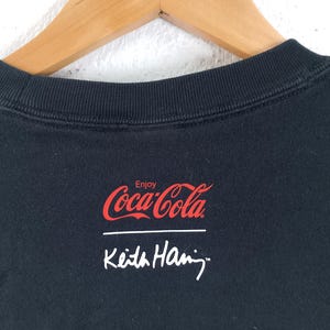 Puede incluir: Sudadera negra de cuello redondo con el logotipo de Coca-Cola en rojo y la palabra "Enjoy" encima. Debajo est&aacute; la firma blanca de Keith Haring. La sudadera est&aacute; colgada en una percha de madera.