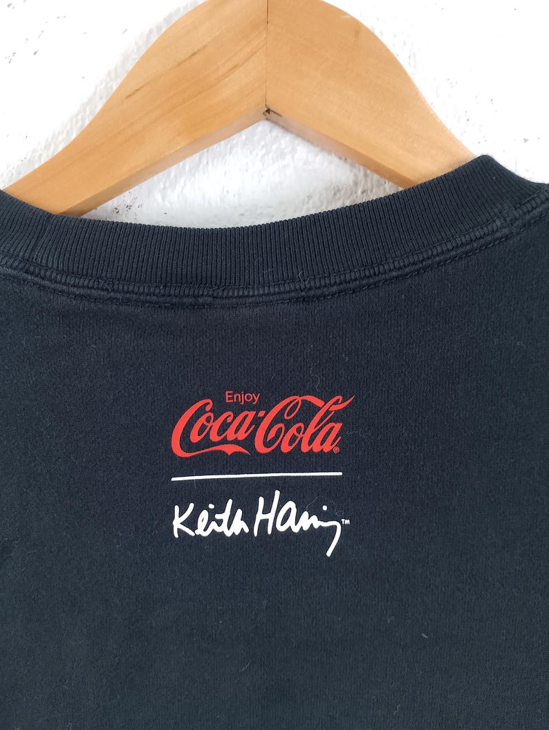 Puede incluir: Sudadera negra de cuello redondo con el logotipo de Coca-Cola en rojo y la palabra "Enjoy" encima. Debajo est&aacute; la firma blanca de Keith Haring. La sudadera est&aacute; colgada en una percha de madera.