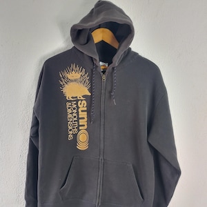 May include: A charcoal gray zip-up hoodie featuring a gold graphic on the chest. The design includes a spiky creature wearing a crown above the text "sunn O))) MONOLITHS & DIMENSIONS". The garment has a drawstring hood and two front pockets.
