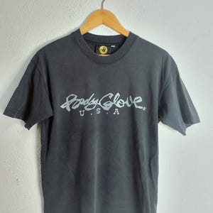 Puede incluir: Una camiseta gris oscuro con la inscripci&oacute;n blanca "Body Glove U.S.A." en una fuente estilizada en la parte delantera. La camiseta est&aacute; colgada de una percha de madera contra una pared blanca lisa. El logotipo de Body Glove es visible en el cuello.