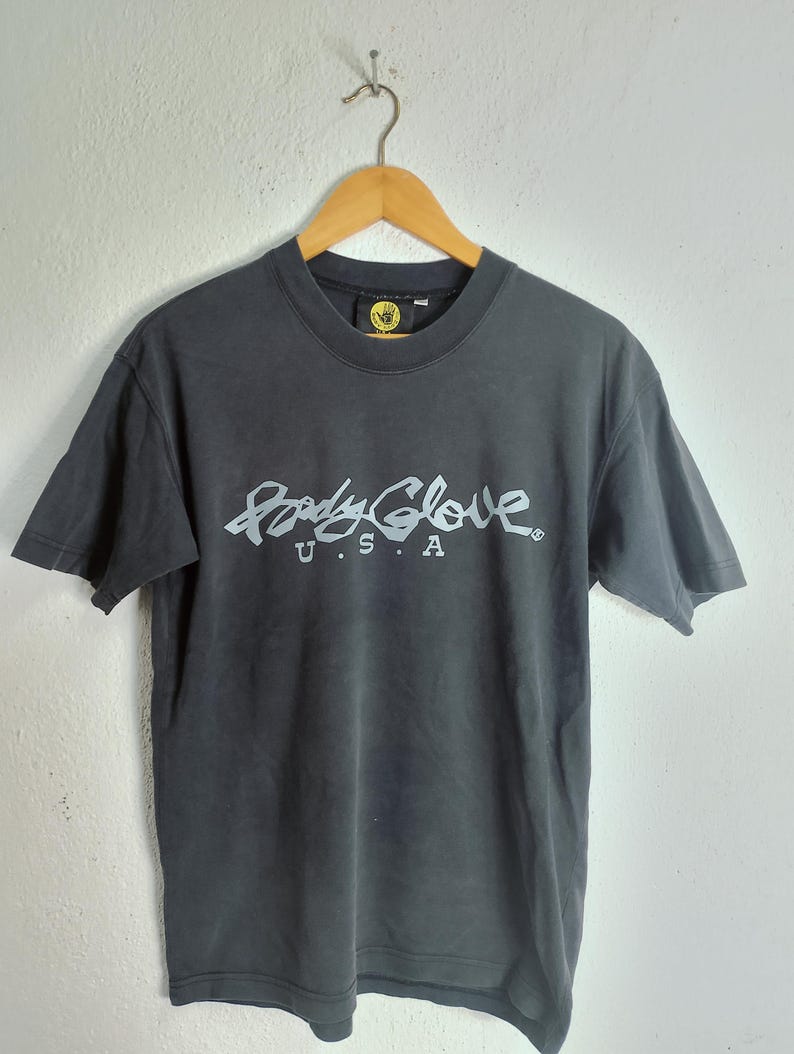 Puede incluir: Una camiseta gris oscuro con la inscripci&oacute;n blanca "Body Glove U.S.A." en una fuente estilizada en la parte delantera. La camiseta est&aacute; colgada de una percha de madera contra una pared blanca lisa. El logotipo de Body Glove es visible en el cuello.