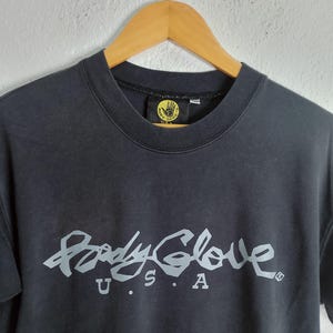 Puede incluir: Camiseta gris oscuro de cuello redondo con el logotipo de Body Glove en gris claro en el pecho. El logotipo incluye las palabras "Body Glove" en una fuente estilizada, con "U.S.A." debajo. La camiseta est&aacute; colgada en una percha de madera.
