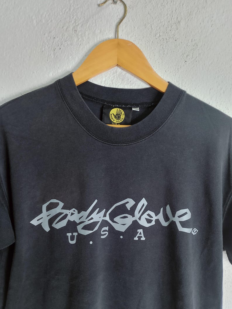 Puede incluir: Camiseta gris oscuro de cuello redondo con el logotipo de Body Glove en gris claro en el pecho. El logotipo incluye las palabras "Body Glove" en una fuente estilizada, con "U.S.A." debajo. La camiseta est&aacute; colgada en una percha de madera.