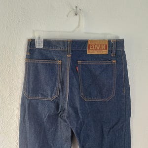 Puede incluir: Vaqueros de mezclilla azul oscuro con dos bolsillos traseros y una peque&ntilde;a etiqueta roja. Los vaqueros tienen un parche de cuero marr&oacute;n con la marca "EDWIN". Las costuras son de color marr&oacute;n claro contrastante.