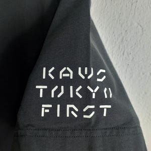 Puede incluir: Manga de camiseta gris oscuro con el texto blanco "KAWS TOKYO FIRST". El texto est&aacute; dispuesto verticalmente. La tela parece ser suave.