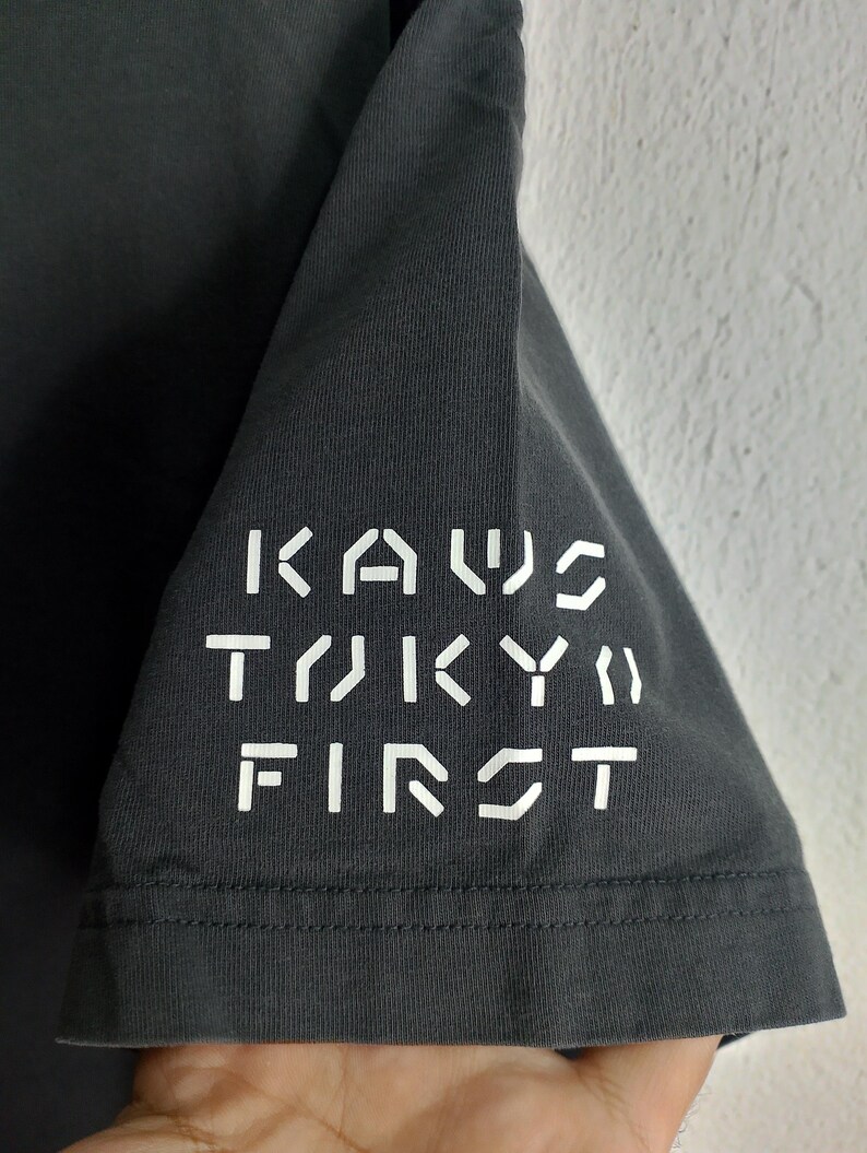 Puede incluir: Manga de camiseta gris oscuro con el texto blanco "KAWS TOKYO FIRST". El texto est&aacute; dispuesto verticalmente. La tela parece ser suave.