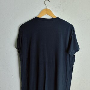 May include: A navy blue t-shirt hanging on a wooden hanger. The t-shirt is made of a soft material and has a simple, classic design. The shirt is hanging against a plain white wall.