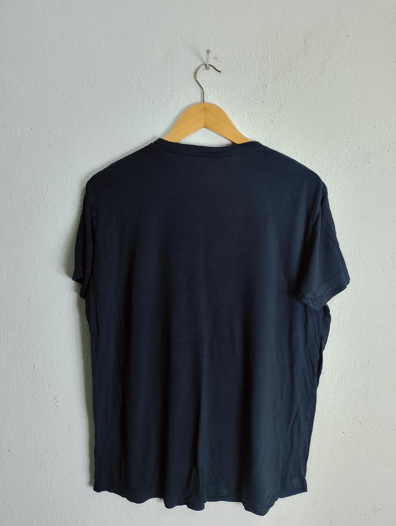 May include: A navy blue t-shirt hanging on a wooden hanger. The t-shirt is made of a soft material and has a simple, classic design. The shirt is hanging against a plain white wall.