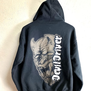 Puede incluir: Sudadera con capucha negra con un gran gráfico de la cara de un diablo en la espalda. La cara del diablo está en tonos grises y marrones, con las palabras "Devil Driver" impresas verticalmente en blanco.
