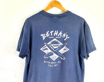 May include: A blue t-shirt with a white graphic on the back. The graphic features the text "BETHANY SURF SHOP" above a diamond shape with crossed surfboards and the text "BETHANY BEACH, DEL. SINCE 1980". The shirt is hanging on a wooden hanger.