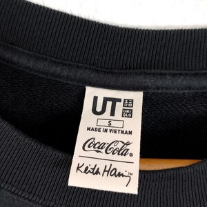 Puede incluir: Sudadera negra de cuello redondo con una etiqueta con el logo de Coca-Cola y la firma de Keith Haring. La etiqueta tambi&eacute;n incluye los logotipos de UT y UNIQLO, la talla S y "Made in Vietnam".