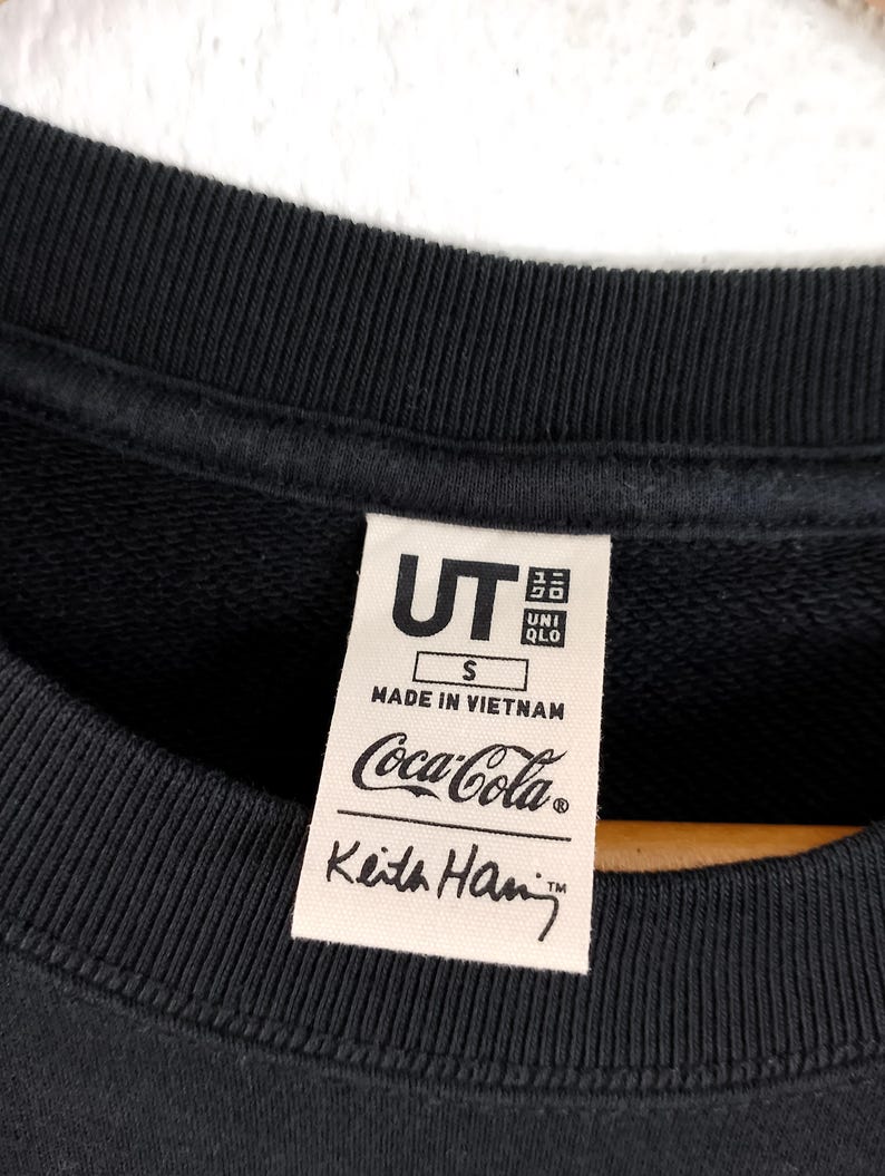 Puede incluir: Sudadera negra de cuello redondo con una etiqueta con el logo de Coca-Cola y la firma de Keith Haring. La etiqueta tambi&eacute;n incluye los logotipos de UT y UNIQLO, la talla S y "Made in Vietnam".