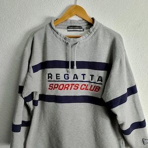 Puede incluir: Una sudadera gris con el texto "REGATTA SPORTS CLUB" en azul marino y rojo. La sudadera tiene rayas azul marino en el pecho y en las mangas. Está colgada en una percha de madera.