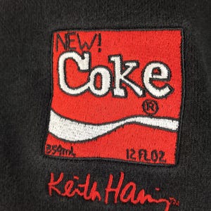 Puede incluir: Sudadera negra con un logotipo "Coke" rojo y blanco bordado, con "NEW!" encima. El logotipo incluye "850ml" y "12 fl oz". El nombre "Keith Haring" est&aacute; bordado en rojo debajo.