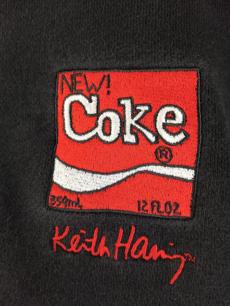Puede incluir: Sudadera negra con un logotipo "Coke" rojo y blanco bordado, con "NEW!" encima. El logotipo incluye "850ml" y "12 fl oz". El nombre "Keith Haring" est&aacute; bordado en rojo debajo.