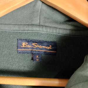 May include: Close-up of a green hoodie with a tag that reads "Ben Sherman." The tag is navy blue with gold lettering and a size indicator. The hoodie is hanging on a wooden hanger.
