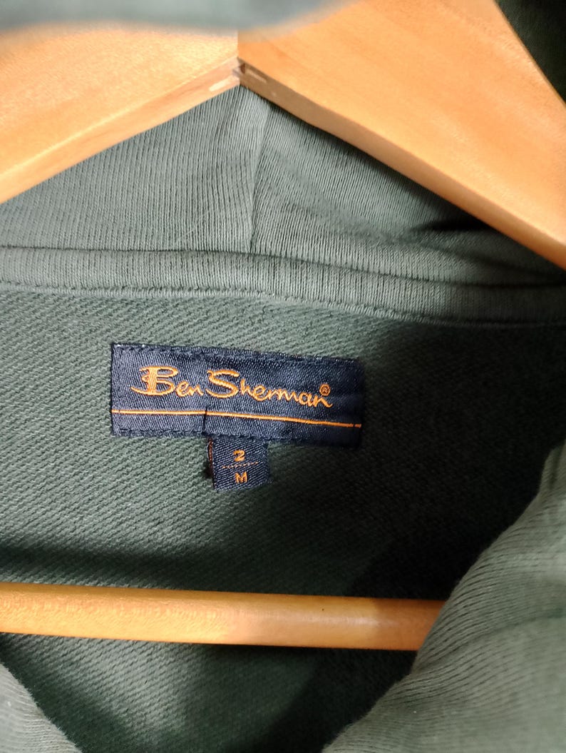 May include: Close-up of a green hoodie with a tag that reads "Ben Sherman." The tag is navy blue with gold lettering and a size indicator. The hoodie is hanging on a wooden hanger.