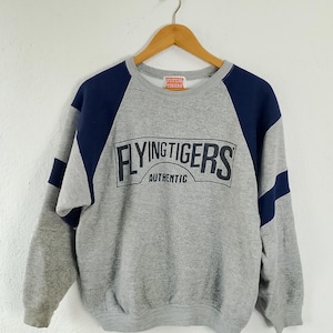 May include: A gray sweatshirt with navy blue shoulder and sleeve accents. The front features the text "FLYING TIGERS AUTHENTIC" in a black, rectangular design. The sweatshirt is hanging on a wooden hanger against a white background.
