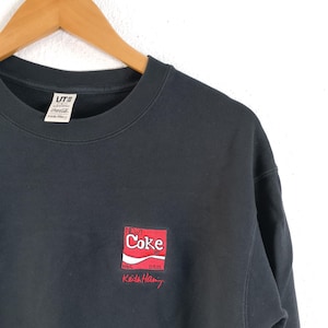 Puede incluir: Sudadera negra de cuello redondo con un peque&ntilde;o dise&ntilde;o bordado de una lata de New Coke en rojo y blanco, con la palabra "Coke". La sudadera tambi&eacute;n tiene la firma "Keith Haring" en rojo. La etiqueta de la marca UT es visible.