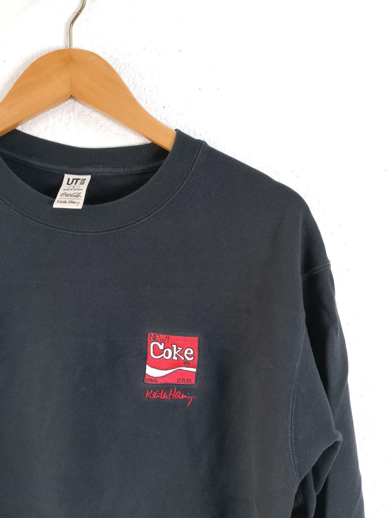 Puede incluir: Sudadera negra de cuello redondo con un peque&ntilde;o dise&ntilde;o bordado de una lata de New Coke en rojo y blanco, con la palabra "Coke". La sudadera tambi&eacute;n tiene la firma "Keith Haring" en rojo. La etiqueta de la marca UT es visible.