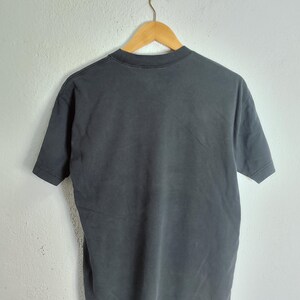 Puede incluir: Una camiseta gris carb&oacute;n colgada de una percha de madera. La camiseta de manga corta es de un material suave y tiene cuello redondo. La camiseta se muestra contra una pared blanca.