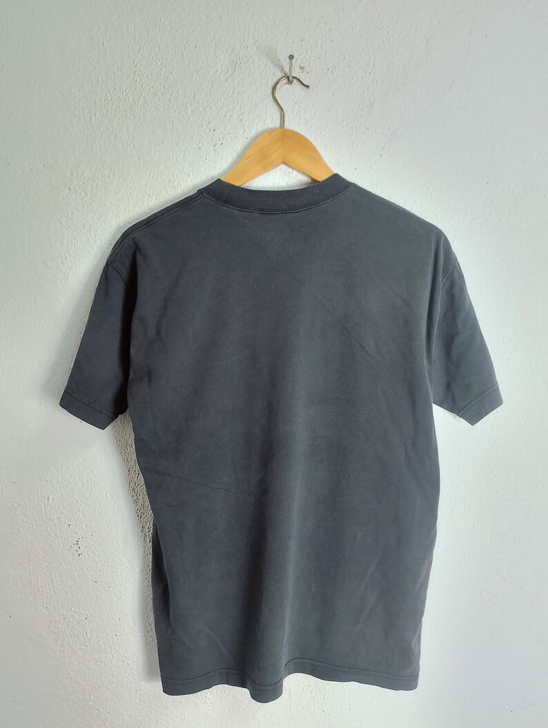 Puede incluir: Una camiseta gris carb&oacute;n colgada de una percha de madera. La camiseta de manga corta es de un material suave y tiene cuello redondo. La camiseta se muestra contra una pared blanca.
