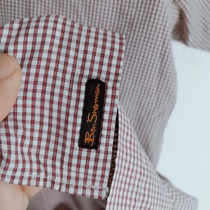K&ouml;nnte beinhalten: Ein kurz&auml;rmeliges Hemd mit einem rot-wei&szlig;en Gingham-Karomuster. Am &Auml;rmelaufschlag befindet sich ein schwarzes, rechteckiges Stoffetikett mit dem in orangefarbenem Garn gestickten Markennamen Ben Sherman. Der Stoff hat eine feine Webstruktur.