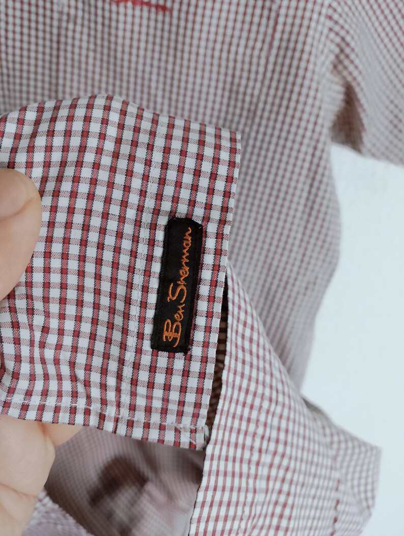 K&ouml;nnte beinhalten: Ein kurz&auml;rmeliges Hemd mit einem rot-wei&szlig;en Gingham-Karomuster. Am &Auml;rmelaufschlag befindet sich ein schwarzes, rechteckiges Stoffetikett mit dem in orangefarbenem Garn gestickten Markennamen Ben Sherman. Der Stoff hat eine feine Webstruktur.