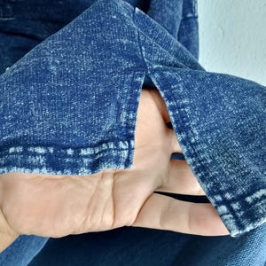 Pode incluir: Close-up de uma pe&ccedil;a de roupa de ganga azul com um aspeto texturizado e desbotado. O tecido tem um efeito desbotado, com &aacute;reas mais claras criando um padr&atilde;o manchado. A borda da pe&ccedil;a de roupa &eacute; vis&iacute;vel, mostrando os detalhes da costura.