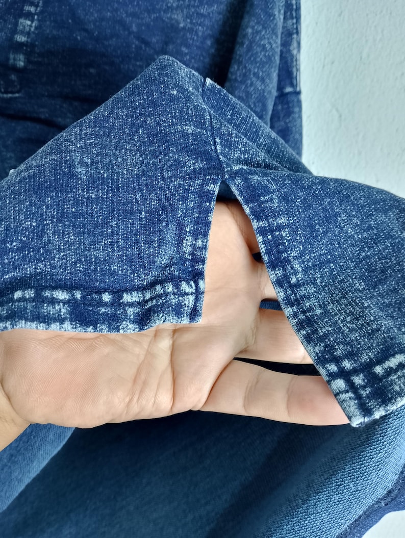 Pode incluir: Close-up de uma pe&ccedil;a de roupa de ganga azul com um aspeto texturizado e desbotado. O tecido tem um efeito desbotado, com &aacute;reas mais claras criando um padr&atilde;o manchado. A borda da pe&ccedil;a de roupa &eacute; vis&iacute;vel, mostrando os detalhes da costura.
