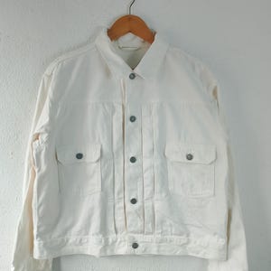 May include: Off-white denim jacket with a classic design. Features a collared neckline, button-down front, two chest pockets, and two side pockets. The jacket is hanging on a wooden hanger against a white wall.