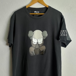 Puede incluir: Camiseta gris oscuro con un gr&aacute;fico de KAWS en la parte delantera. El gr&aacute;fico representa una figura sentada con las manos cubriendo su rostro. La manga derecha tiene el texto "KAW TOKYO FIRST". La camiseta est&aacute; colgada en una percha de madera.