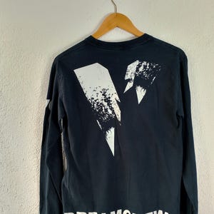 Pode incluir: Camiseta preta de manga comprida com um design gr&aacute;fico branco nas costas. O design inclui um raio e o texto "DREAMCREW" em letras brancas. A camiseta est&aacute; pendurada em um cabide de madeira.